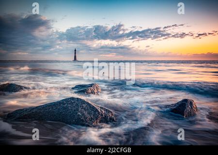 Ocean tide and rocks with lighthouse in the distance seen during colorful sunrise Stock Photo
