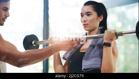 Dedication is key. Cropped shot of an attractive young sportswoman lifting a barbell with the help of her fitness coach in a gym. Stock Photo