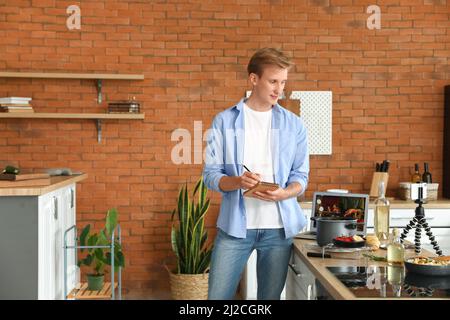 Young man making notes while watching video tutorial in kitchen Stock Photo