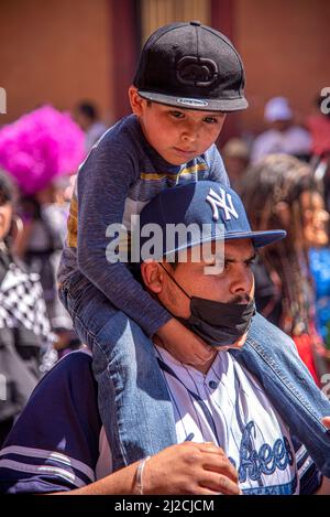 A Mexican father wearing a mask, carrying his son who is wearing a cap on his shoulders. San Miguel de Allende, Guanajuato, Mexico Stock Photo