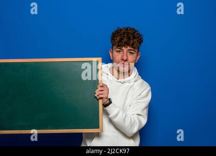 Caucasian young man with curly hair holding a blackboard isolated on blue studio background. Learning and messaging concept. Stock Photo