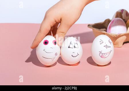 Children's hand lays painted Easter white eggs on a pink and white background. Funny and romantic faces drawn with a marker. The concept of children's needlework. Copy space. Stock Photo