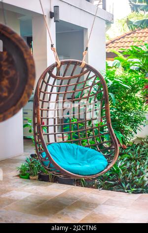 Wicker hanging cozy chair with a pillow on the site near the house, next to the tropical garden. Decor and landscape design. Stock Photo