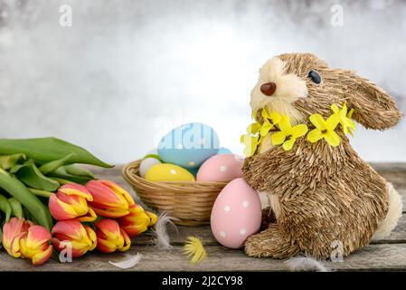 Funny bunny made of straw with Easter eggs painted, feathers and orange-yellow tulips on wooden background. Easter concept. Stock Photo