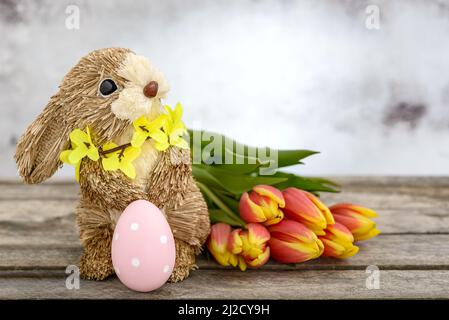 Funny bunny made of straw with Easter egg painted and orange-yellow tulips on wooden background. Easter concept. Stock Photo