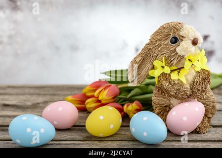 Funny bunny made of straw with Easter eggs painted and orange-yellow tulips on wooden background. Easter concept. Stock Photo