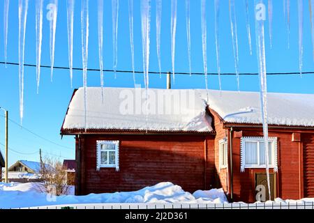 Long transparent icicles hang on roof of house in village in northern Europe at winter sunny day. Stock Photo