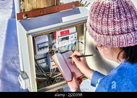 Woman is taking readings from external electric meter and writing them into notebook outside in winter. Stock Photo