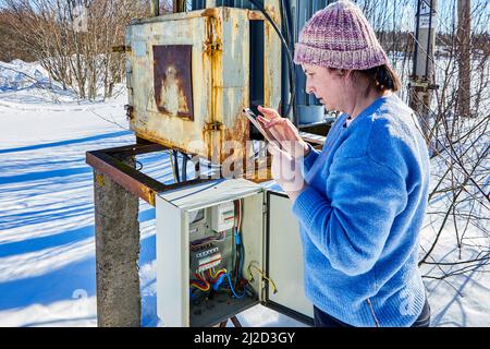 Checking readings from external electric meter outdoors by woman with smartphone in countryside. Stock Photo