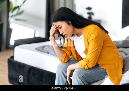 Upset sad Chinese brunette young woman, in casual clothes, sitting on the bed at home, saddened by personal problems, news, loneliness, experiencing stress, uncertainty Stock Photo