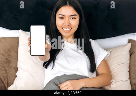 Gadgets and technology, mock-up concept. Happy pretty asian young woman in basic white t-shirt, showing her smart phone with empty white mock-up screen while lying in bed at home, smiling Stock Photo