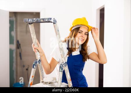 Smiling female builder in blue overalls and yellow hard hat at construction site Stock Photo