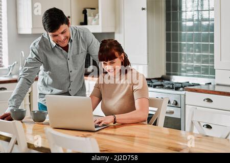 Hows your online course going, babe. Shot of a woman using a laptop with her husband standing beside her. Stock Photo