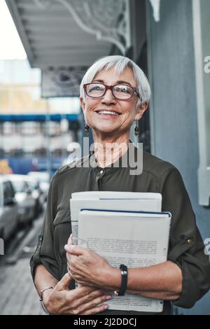 This city always reminds me of my youth. Portrait of a cheerful mature businesswoman carrying books and paperwork while out in the city. Stock Photo