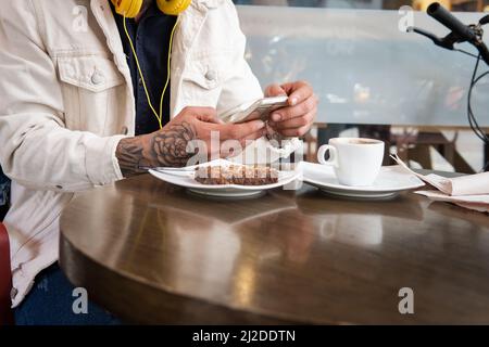 Close-up of the hands of an unrecognizable man who is typing in a message on his cell phone or smartphone at a table in a coffee shop next to a cup of Stock Photo