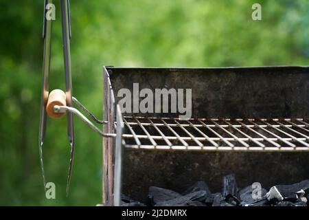Barbecue tongs (Grillzange) on a wooden handle from a charcoal grill with dirty rust. Green nature in the background. Stock Photo