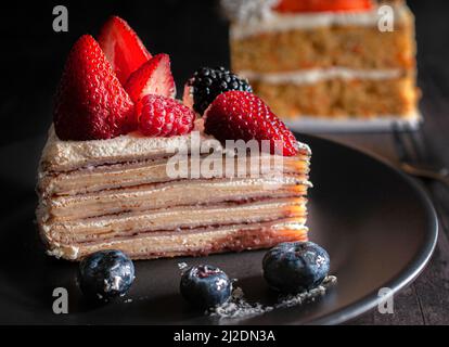 Slice of banana crepe cake with cream and strawberries on top with blue berries on a black plate. macro, dark food photography. Stock Photo