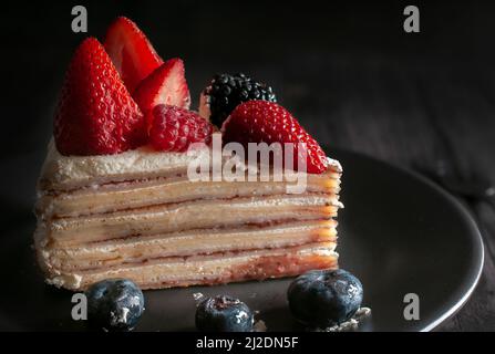 Slice of banana crepe cake with cream and strawberries on top with blue berries on a black plate. macro, dark food photography. Stock Photo