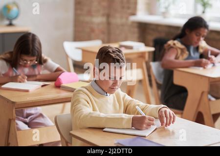 High angle portrait of cute schoolboy sitting at desk in classroom with wooden decor, copy space Stock Photo