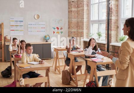 Diverse group of children raising hands in school classroom with young female teacher speaking, copy space Stock Photo