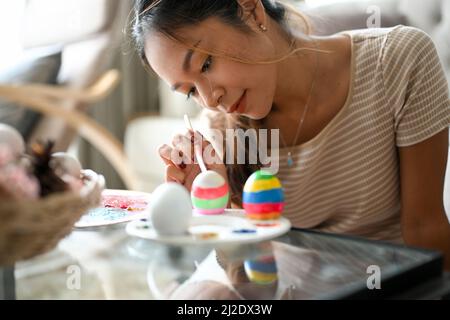 Relaxed and clam woman painting on an Easter egg with a colourful colour in her home living room. Happy Easter holiday concept. Stock Photo