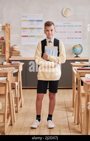 Vertical full length portrait of smiling boy in school looking at camera while standing in classroom Stock Photo