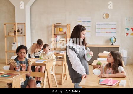 Diverse group of children chatting in school classroom during break, focus on two girls enjoying lunch in foreground, copy space Stock Photo