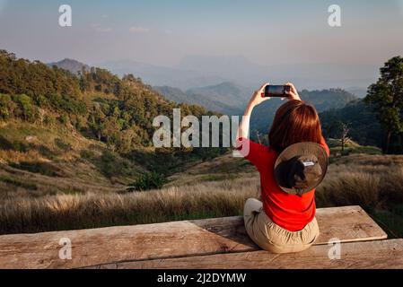A woman in a red long-sleeve with a hat sits on the wooden balcony and takes a photo of the golden dry grasslands at Hadubi, Chiang Mai, Thailand. Stock Photo