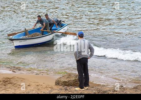 Fishermen standing inside a boat holding paddles going out of the water, full of fish trapped in the fishing net. A man looking at them. Stock Photo
