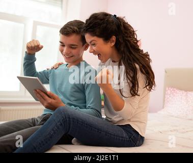 Mother and son feeling happy while sitting in a bedroom and playing an video game on tablet. Stock Photo