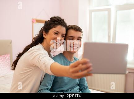 Smiling mother and son sitting at bedroom using a tablet to take a photo of themselves. Stock Photo