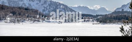 winter landscape with people on snowy frozen lake and touristic mountain village , shot in bright light near Maloja, Grisons, Switzerland Stock Photo
