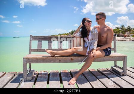 Young couple sits on a bench on a pier next to the turquoise sea, enjoying their summer holiday in tropical destination. Stock Photo