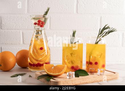 lemonade from orange and berries in two modern glasses in the form of a can and a carafe on a table against a brick wall Stock Photo