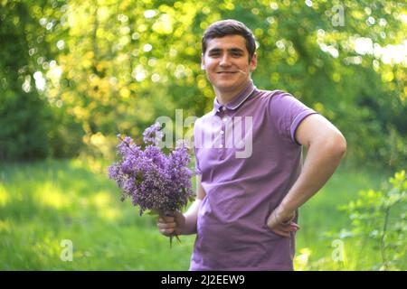 Defocus pleasant cheerful handsome smiling young guy looking aside and holding bouquet of lilac flowers while feeling happy. Young beautiful man with Stock Photo