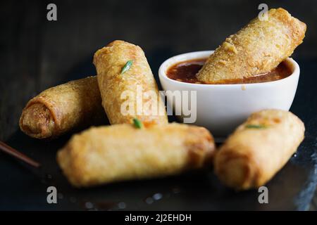 Egg rolls in sweet and sour sauce over a dark rustic table. Garnished with green onions. Selective focus with blurred foreground and background. Stock Photo