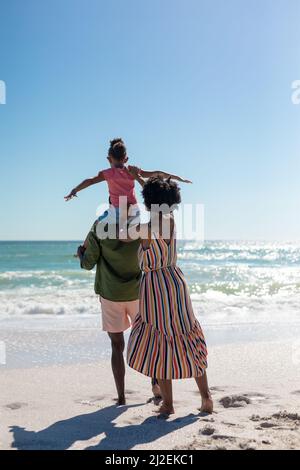 African american woman supporting daughter sitting on her father's shoulders at beach Stock Photo