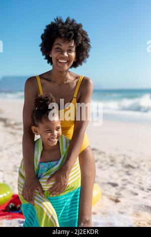 Portrait of happy african american woman with daughter wrapped in towel at beach on sunny day Stock Photo