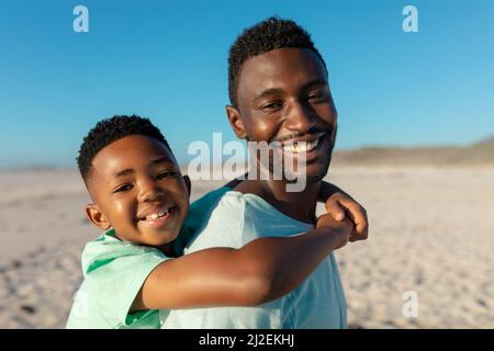 Side view of portrait of smiling african american father piggybacking son at beach on sunny day Stock Photo