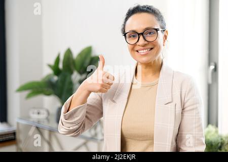 Woman entrepreneur stands and showing big finger in the contemporary office, freelancer woman in smart casual wear looks at the camera with arms crossed, small business owner Stock Photo