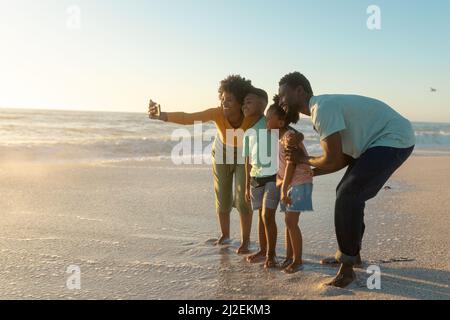 Happy african american mother taking selfie with family on shore at beach during sunset Stock Photo