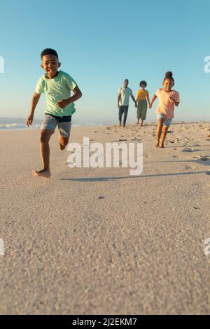Happy african american siblings running against parents walking at beach on sunny day Stock Photo