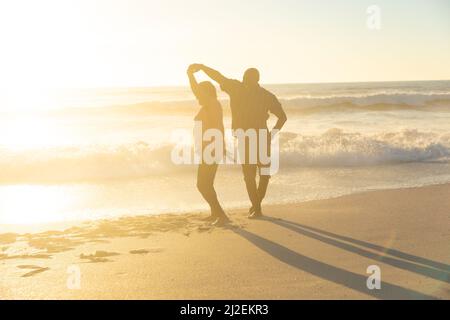 Happy african american senior couple dancing on shore at beach during sunset Stock Photo