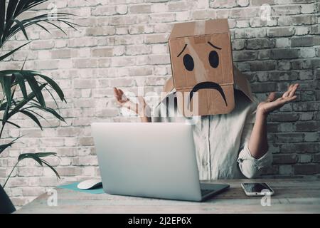 Modern worker with carton sad box on head open arms with desolation gesture in front of a laptop. Online crypto smart working business job activity. C Stock Photo