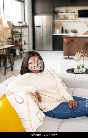 Healthy multiracial woman with sitting on the pure comfortable sofa at modern apartment, dressed in casual clothes. Lady looking at the camera Stock Photo