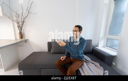 Young man sitting on couch while switching TV channels. Focus on remote control. Stock Photo