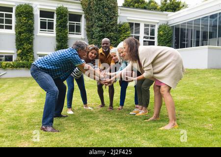 Multiracial senior male and female friends stacking hands while playing rugby in backyard Stock Photo