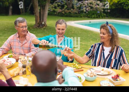 Multiracial senior male and female friends having wine at table during backyard party Stock Photo