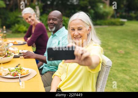 Senior woman taking selfie with multiracial friends having food at backyard party Stock Photo