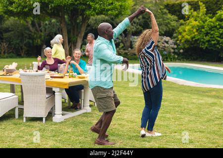 Multiracial senior couple dancing while friends at table in background during backyard party Stock Photo